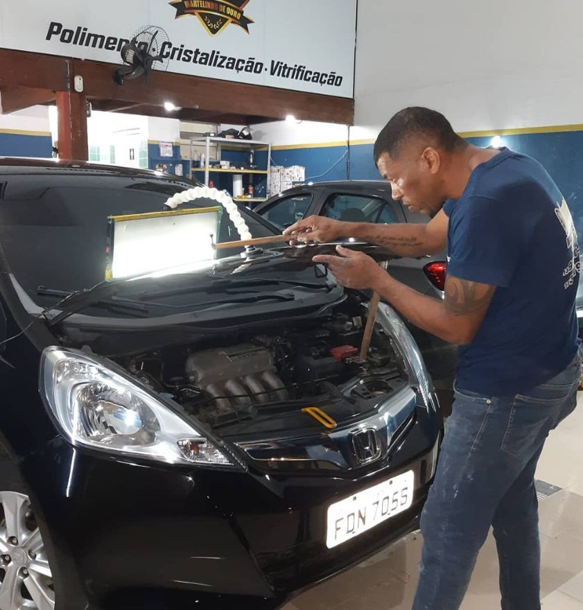 O Bico desamassando o capô de um Honda Fit preto, com a luz de inspeção sobre a lataria