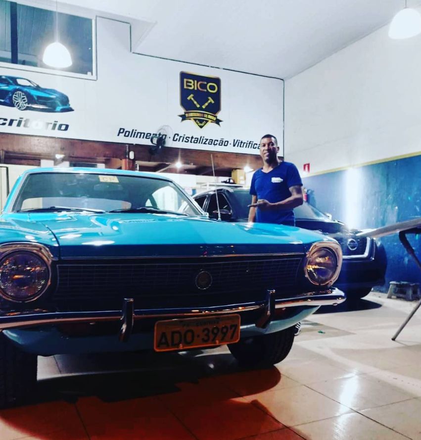 O Bico ao lado de um Ford Maverick azul clássico, dentro da oficina